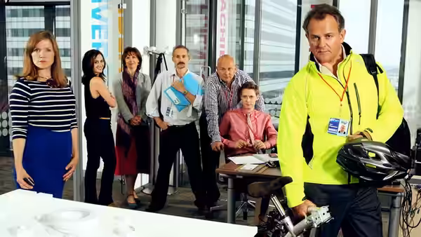 Ian Fletcher (Hugh Bonneville) carries a bicycle helmet through the office as colleagues gather behind him
