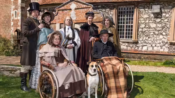 Victorian-dressed villagers gather with a dog outside a stone building