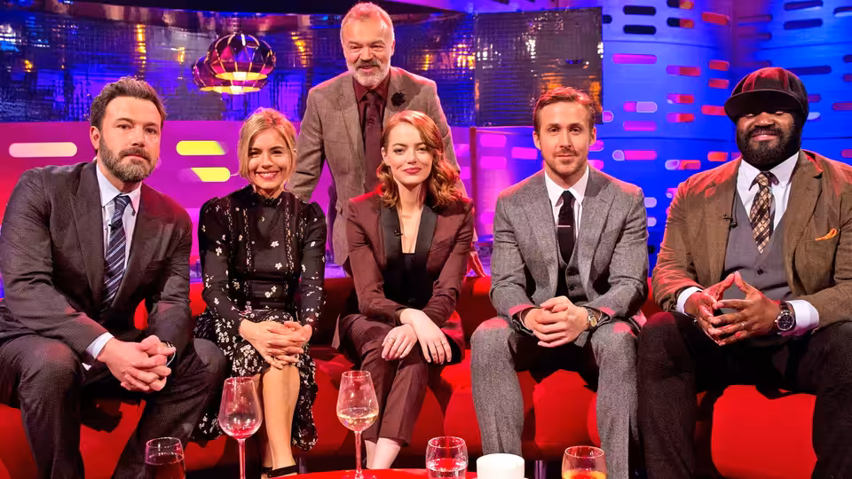 Graham Norton stands behind five guests seated together on the brightly lit studio sofa