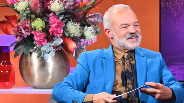 Graham Norton smiles while holding a pen beside a large vase of flowers
