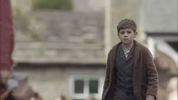 A boy in a brown coat stands outside a stone village house