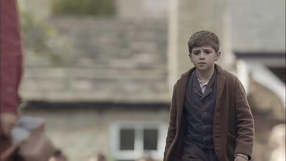 A boy in a brown coat stands outside a stone village house
