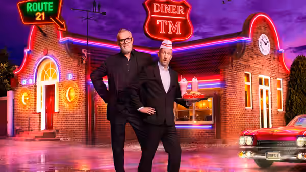 Greg Davies and a suited man in a fez pose outside a neon-lit diner