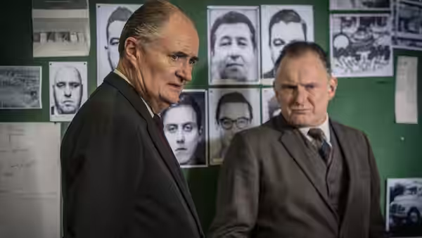 Two detectives examine a wall covered with portraits and investigation materials