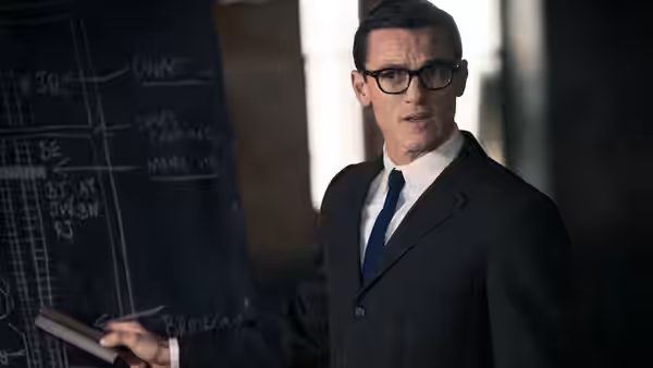 A bespectacled man points to a chalkboard covered with diagrams