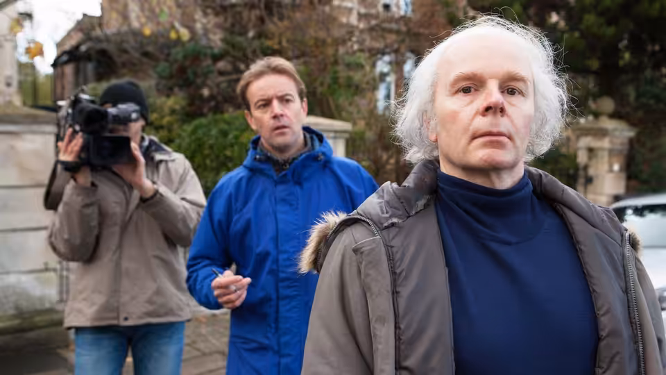 Christopher Jefferies (Jason Watkins) walks past a cameraman as a man in blue follows