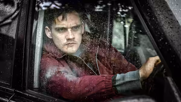 A man in a red jacket sits behind the wheel of a rain-spattered car