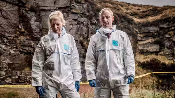 Two investigators in protective suits examine a rocky outdoor crime scene behind police tape