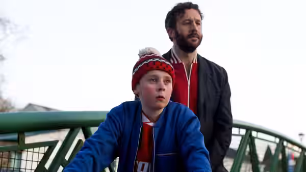 Martin Moone (David Rawle) rides a bicycle across a green metal bridge with Sean Murphy (Chris O'Dowd) behind him