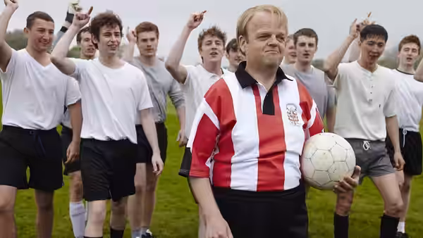 Neil Baldwin (Toby Jones) holds a football among young players on the pitch