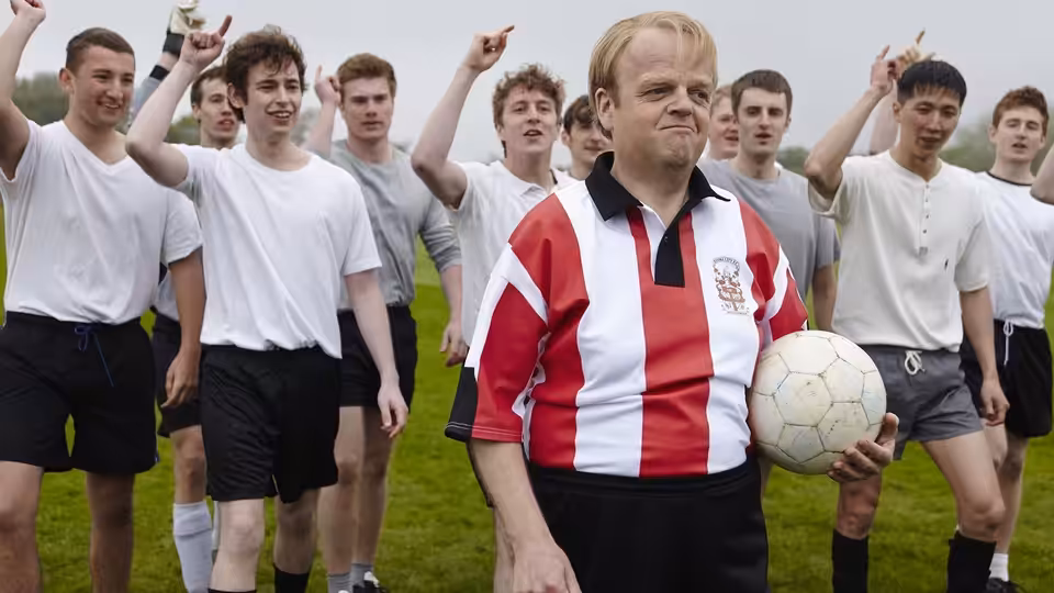 Neil Baldwin (Toby Jones) holds a football among young players on the pitch