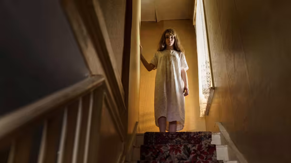 Janet Hodgson (Eleanor Worthington-Cox) stands at the top of a dimly lit staircase