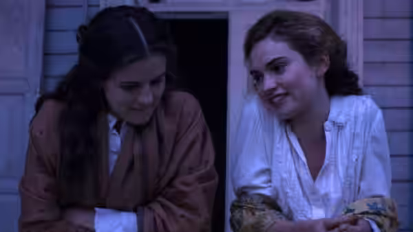 Two young women sit together outside a house, sharing a quiet conversation
