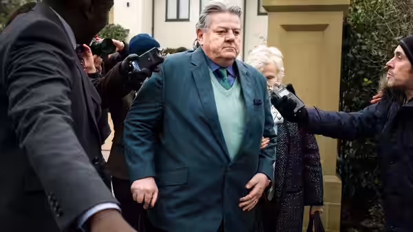 Paul Finchley (Robbie Coltrane) walks outside as reporters extend cameras and microphones toward him