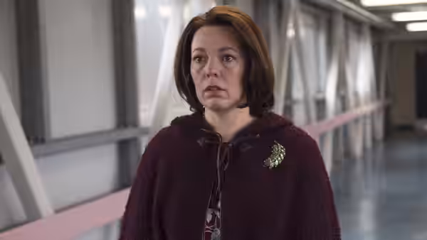 Deborah Flowers (Olivia Colman) stands in a covered walkway wearing a burgundy cape