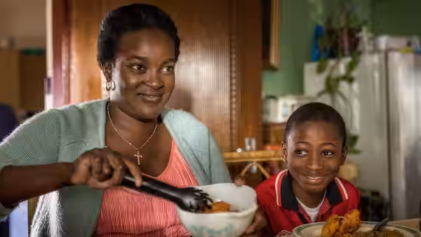 Gloria Taylor (Wunmi Mosaku) serves food beside Damilola (Sammy Kamara) at the kitchen table