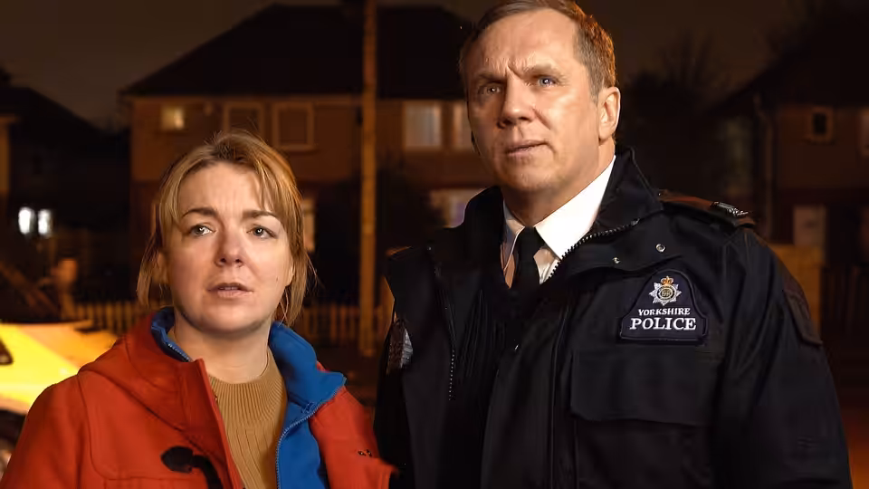 Julie Bushby (Sheridan Smith) stands beside a uniformed police officer outside houses at night