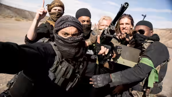 Masked armed men gather outdoors, one raising a finger beside a rifle