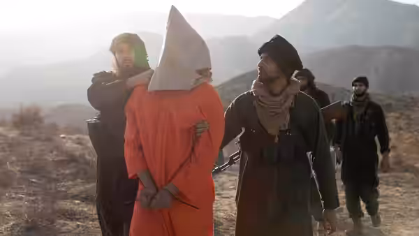 Armed men escort a hooded prisoner in an orange jumpsuit across rocky mountain terrain