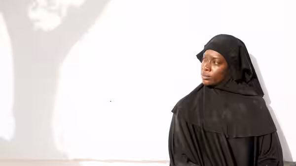A woman in a black veil stands beside a sunlit white wall
