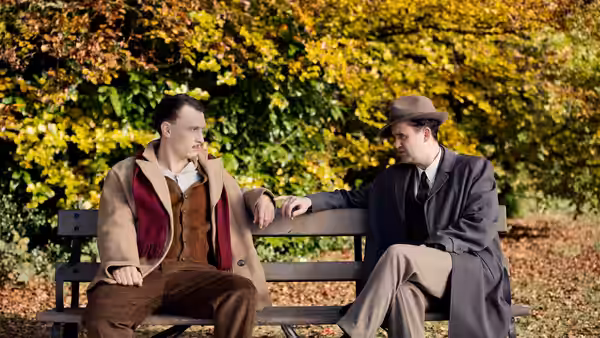 Two men sit talking on a park bench beneath autumn leaves
