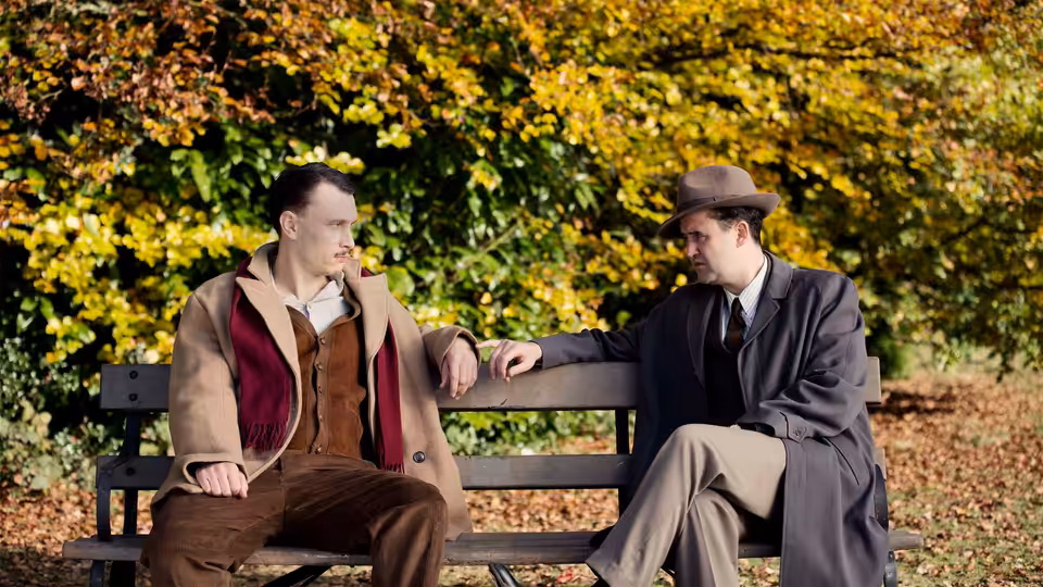 Two men sit talking on a park bench beneath autumn leaves