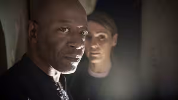Nelly (Lennie James) stands in a doorway while a woman watches from behind