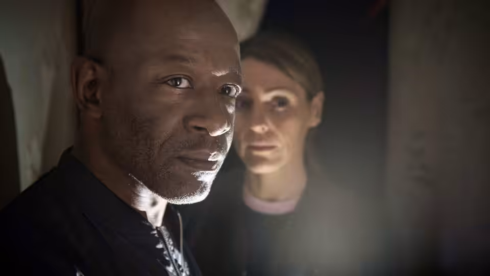 Nelly (Lennie James) stands in a doorway while a woman watches from behind