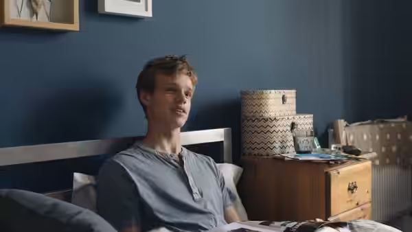 A young man sits up in bed beside a wooden chest of drawers