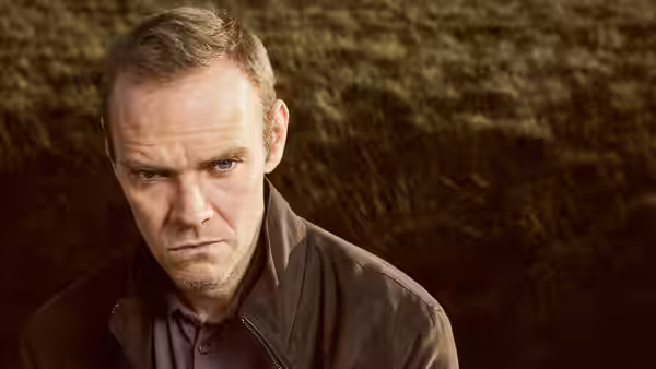 A man in a brown jacket looks ahead from a grassy hillside