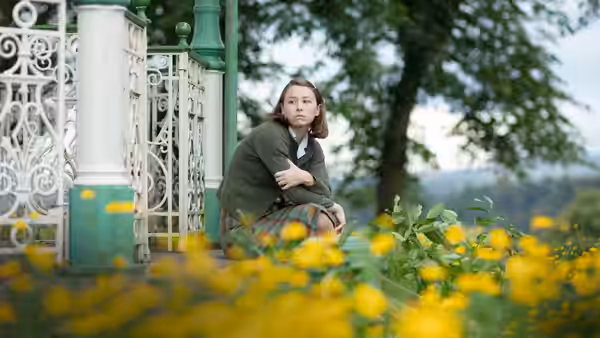 Young Maud Palmer (Liv Hill) crouches beside an ornate garden gate among flowers
