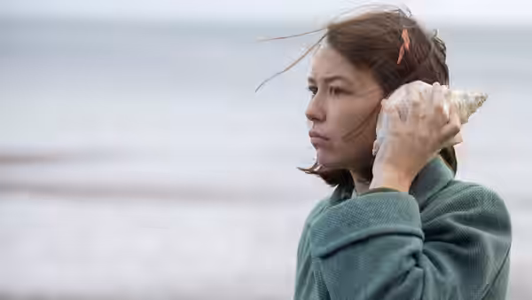 A young woman holds a seashell to her ear beside the sea