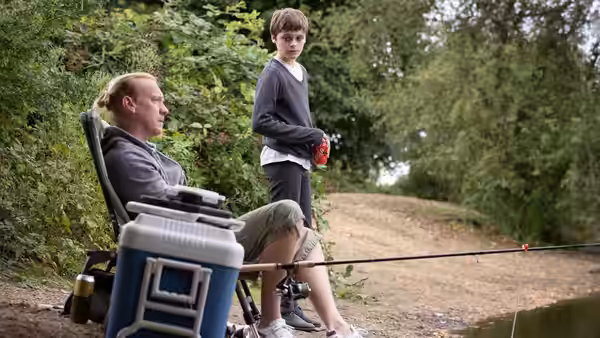 Ray (Billy Barratt) stands beside a seated man fishing beside a wooded path