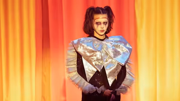 A woman in a metallic clown costume stands before orange stage curtains