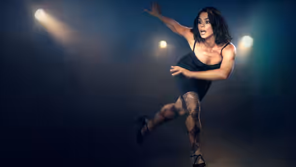 A woman in a black outfit dances beneath spotlights on a dark stage