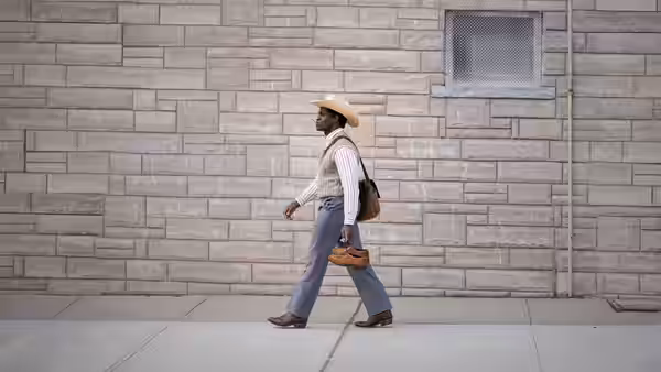 A man in a cowboy hat walks beside a stone wall carrying a bag