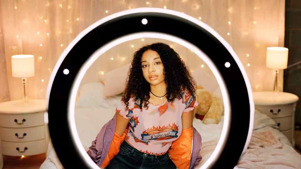 A young woman sits on a bed framed by a large illuminated ring