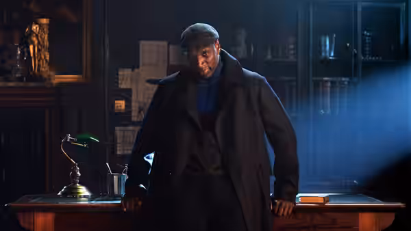 A man in a flat cap leans against a desk in a dim office