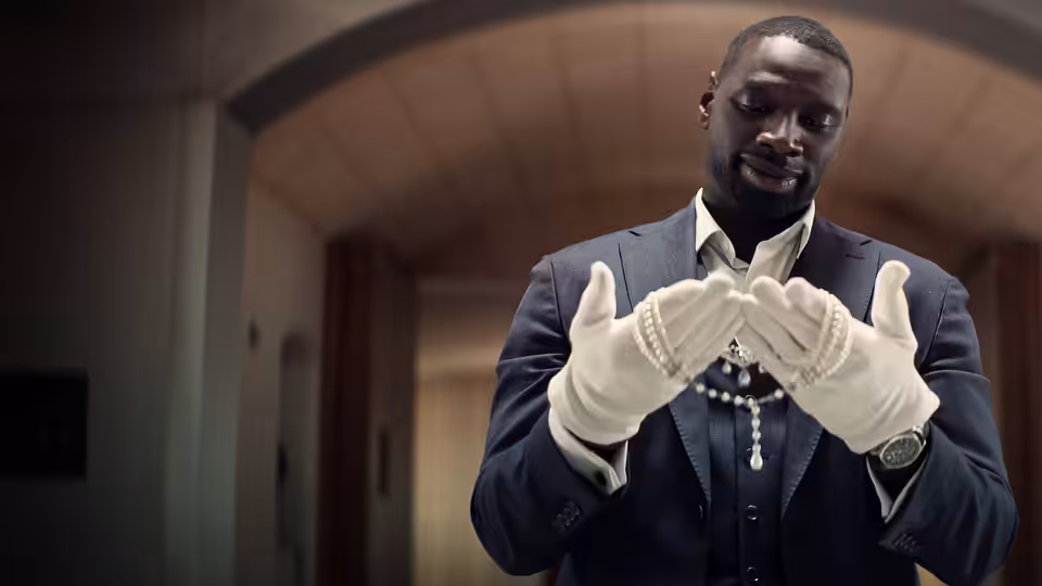 Assane Diop (Omar Sy), dressed as a museum attendant, examines his white gloves indoors