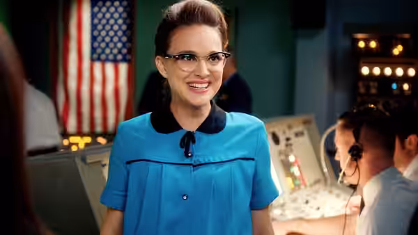 A smiling woman in glasses and a blue uniform stands in a control room