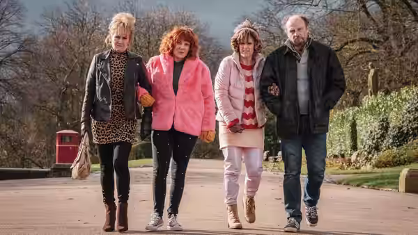 Alma walks arm in arm with a woman and man along a park path