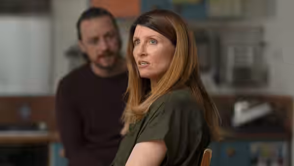She (Sharon Horgan) turns toward someone while He (James McAvoy) stands behind her in a kitchen