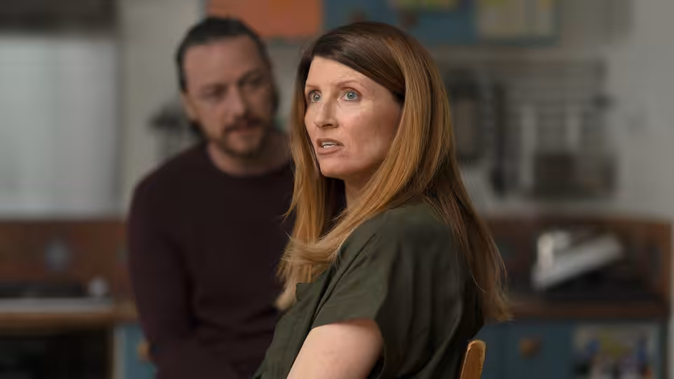 She (Sharon Horgan) turns toward someone while He (James McAvoy) stands behind her in a kitchen