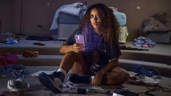 Sasha (Nicôle Lecky) sits amid scattered clothes, holding her phone