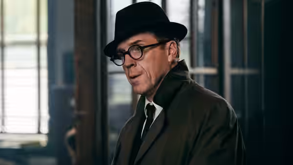 A bespectacled man in a fedora and overcoat stands inside a dim room