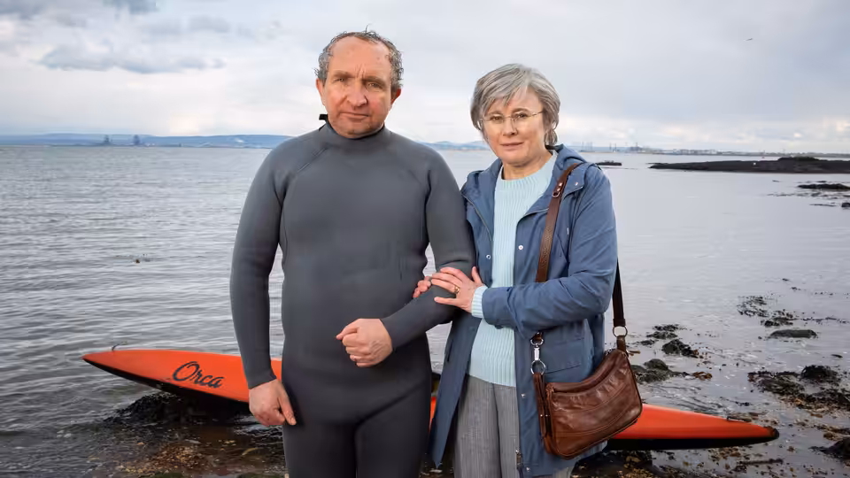 John Darwin (Eddie Marsan) in a wetsuit beside Anne Darwin (Monica Dolan) on the shore by a red canoe
