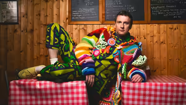 Joe Lycett lounges across café tables in a multicoloured outfit