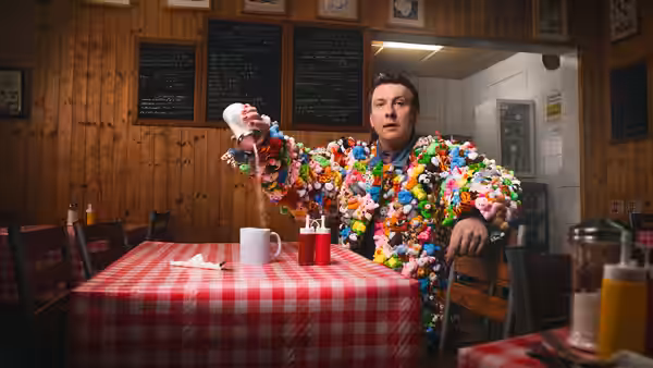 Joe Lycett in a brightly decorated suit sits at a café table, holding a small object