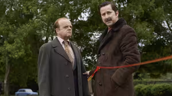 Two men in overcoats stand outdoors beside a red cordon