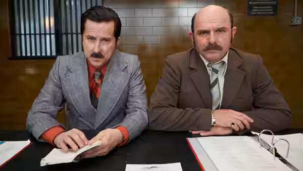Two moustached men in suits sit at a desk, reviewing papers in a tiled office